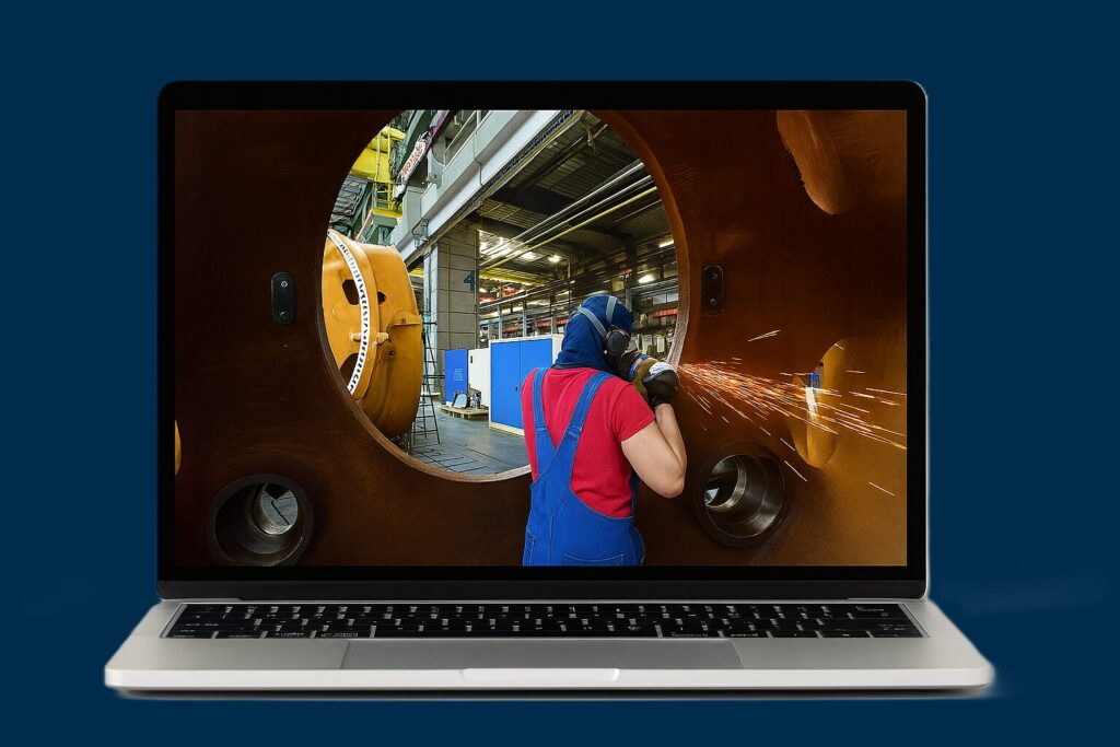 Worker in blue overalls and red shirt welds inside a large circular rust-colored metal component, sparks flying in a factory setting viewed through the opening on a laptop screen. (Informative)
