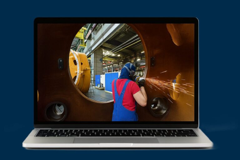 Worker in blue overalls and red shirt welds inside a large circular rust-colored metal component, sparks flying in a factory setting viewed through the opening on a laptop screen. (Informative)